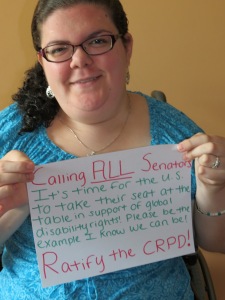 Young caucasian woman with curly brown hair off to the side wearing purple glasses and a turquoise floral-patterned shirt. She is holding up a sign written in pink and green ink on white paper that reads: "Calling all senators: It's time for the U.S. to take their seat at the table in support of global disability rights! Please be the example I know we can be! Ratify the CRPD!"